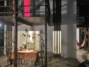 An outdoor dining area with a table and chairs, a hammock, and a view of a building with vertical blinds. - La Réserve Design Villas (Tibau do Sul)