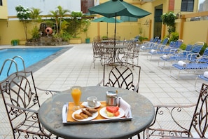A poolside dining area with metal chairs, a table with breakfast items, and a pool in the background.