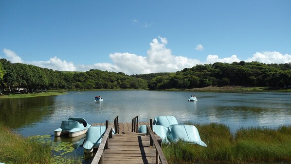 A wooden dock leading into a calm lake with paddle boats and a boat in the distance. - Fiore Resort e Residence (Paripueira)