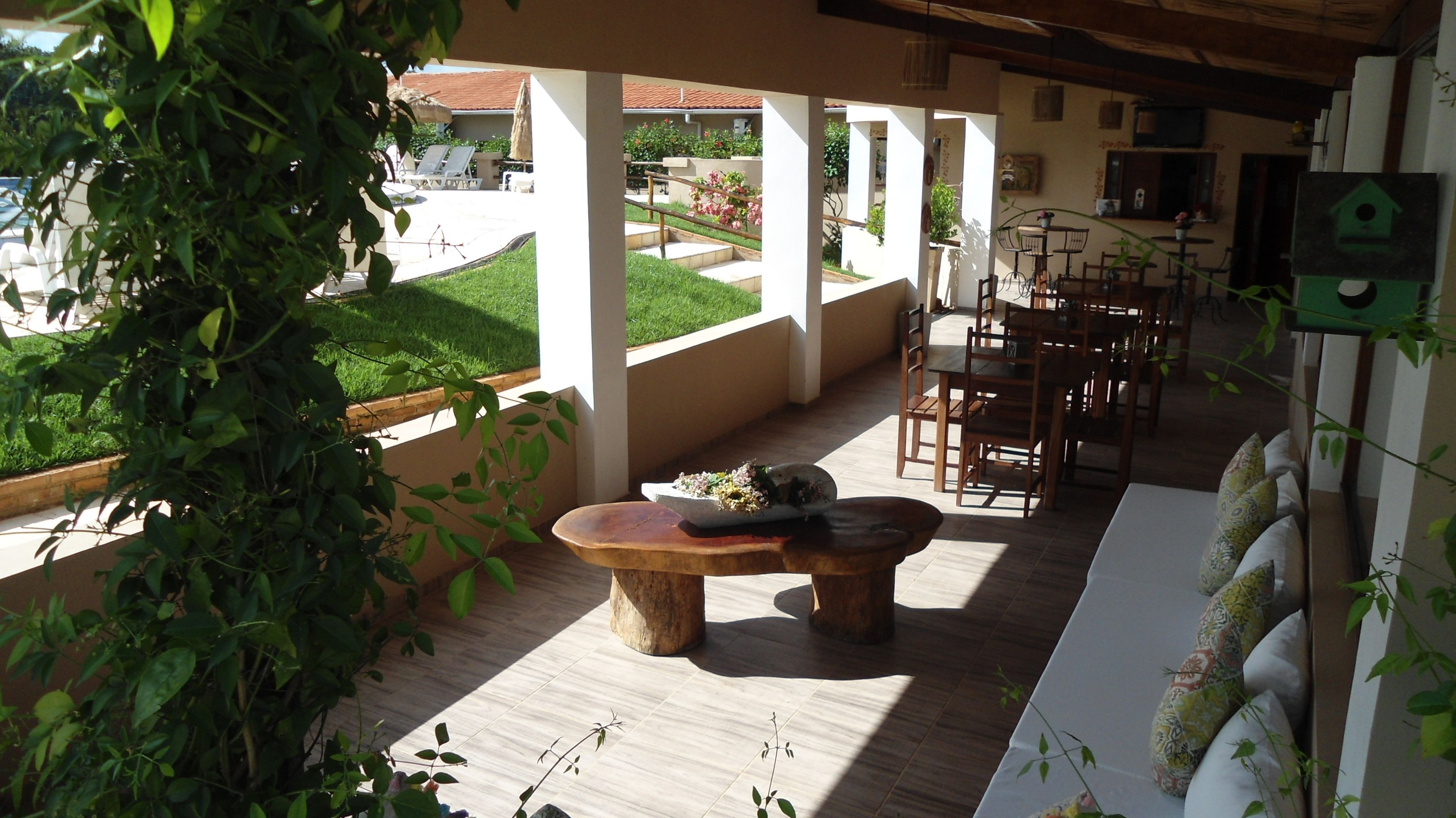 Un espacio al aire libre con techo, mesas y sillas de madera, rodeado de plantas en macetas y vistas a un jardín.