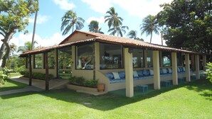 A covered outdoor seating area with a low wall and a tiled roof, surrounded by lush greenery and palm trees. - Fiore Resort e Residence (Paripueira)