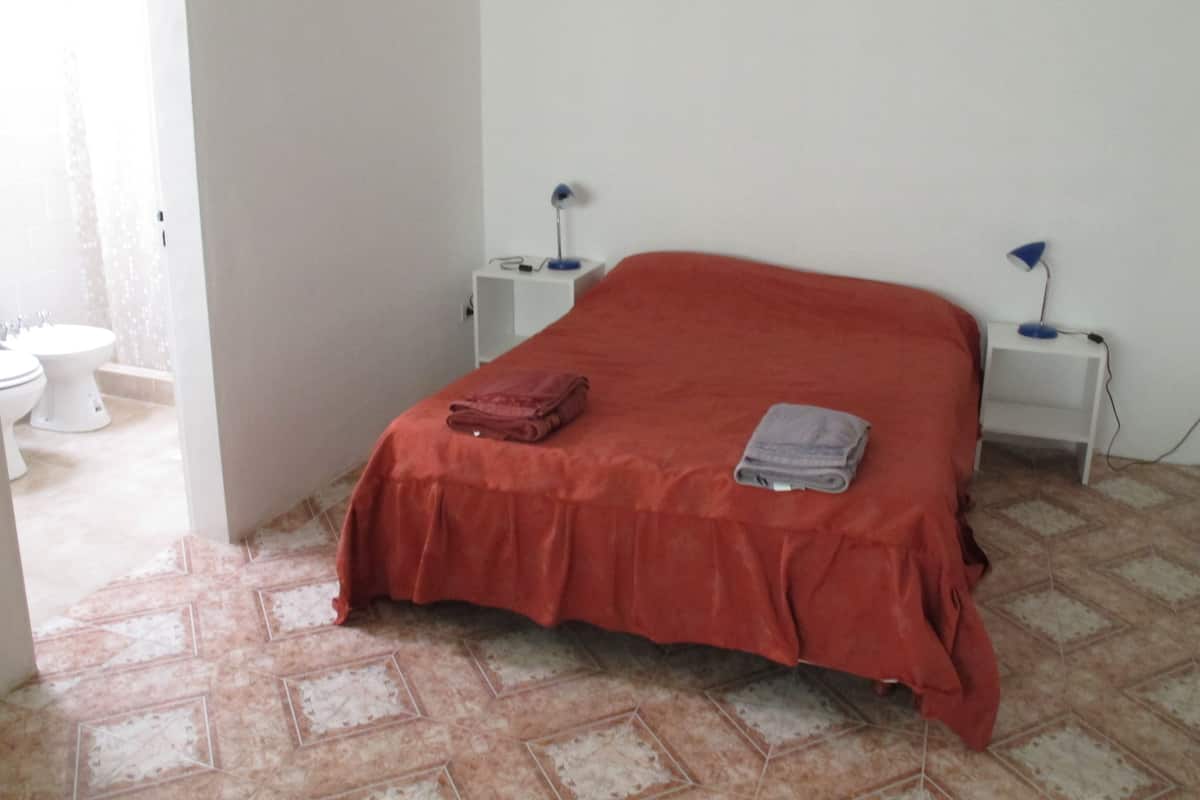 A room with a bed, a nightstand, a lamp, a folded towel, and a toilet.