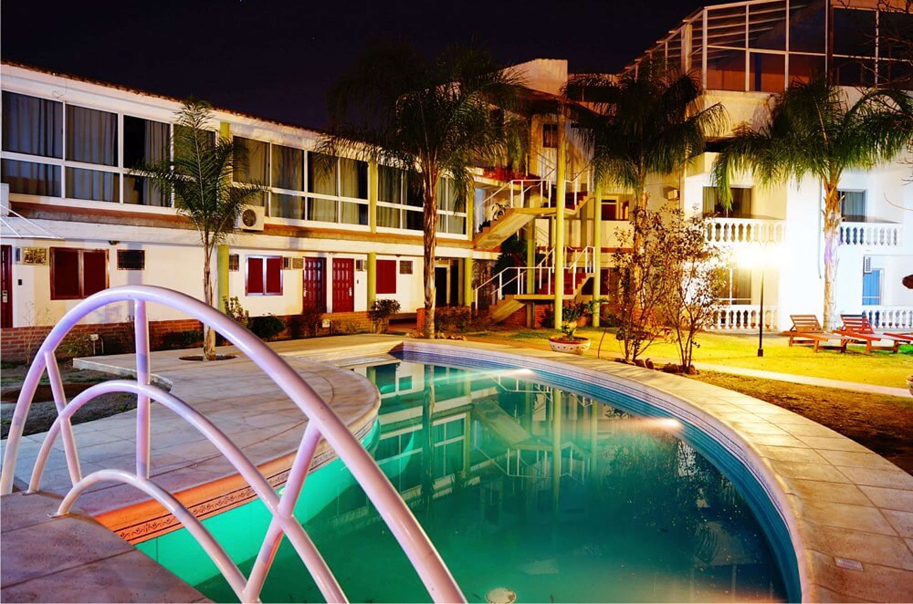 Una piscina con plataforma de metal, rodeada de edificios de varios pisos y balcones, y palmeras.