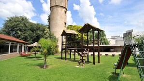 A grassy area with a wooden play structure, a tall cylindrical tower, and a building with a red roof. - Hotel El Cid (Villa Carlos Paz)