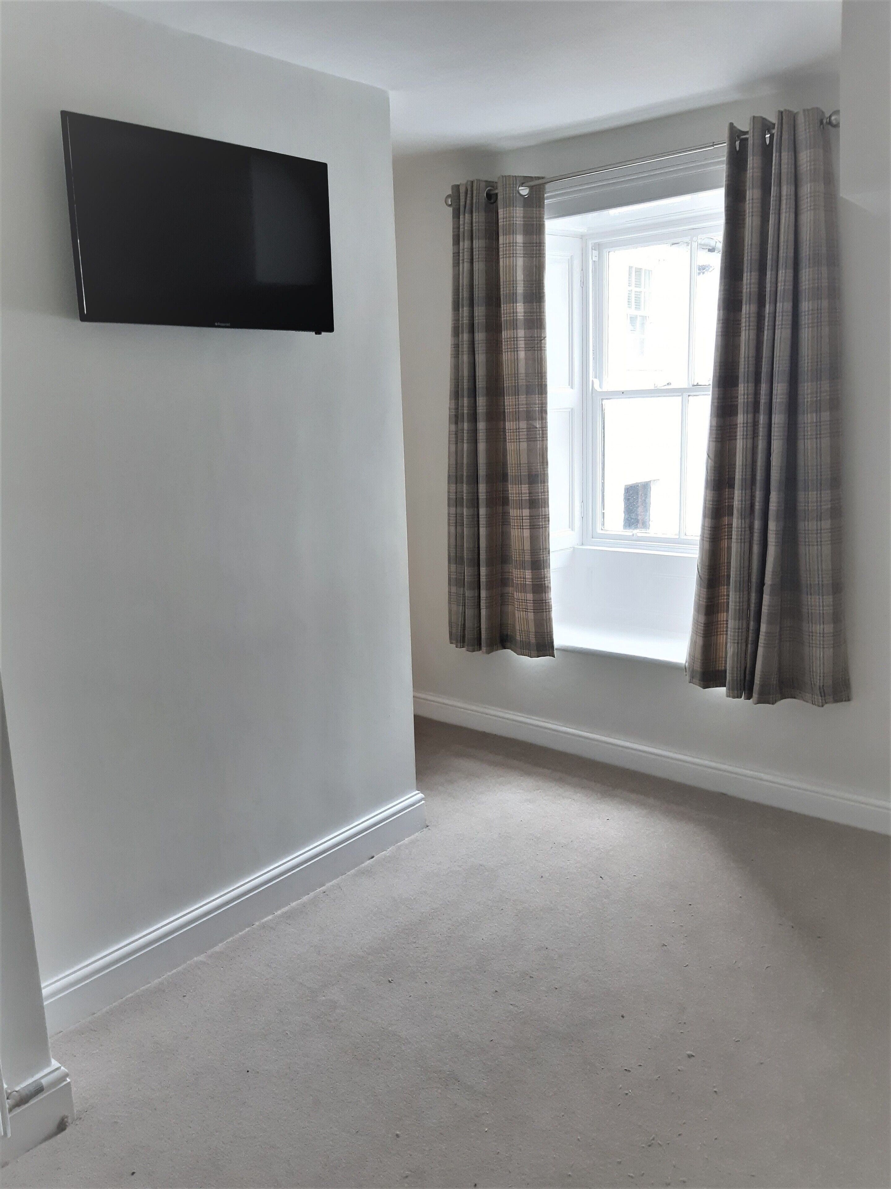 A room with a flat-screen TV mounted on the wall, a window with plaid curtains, and a carpeted floor.