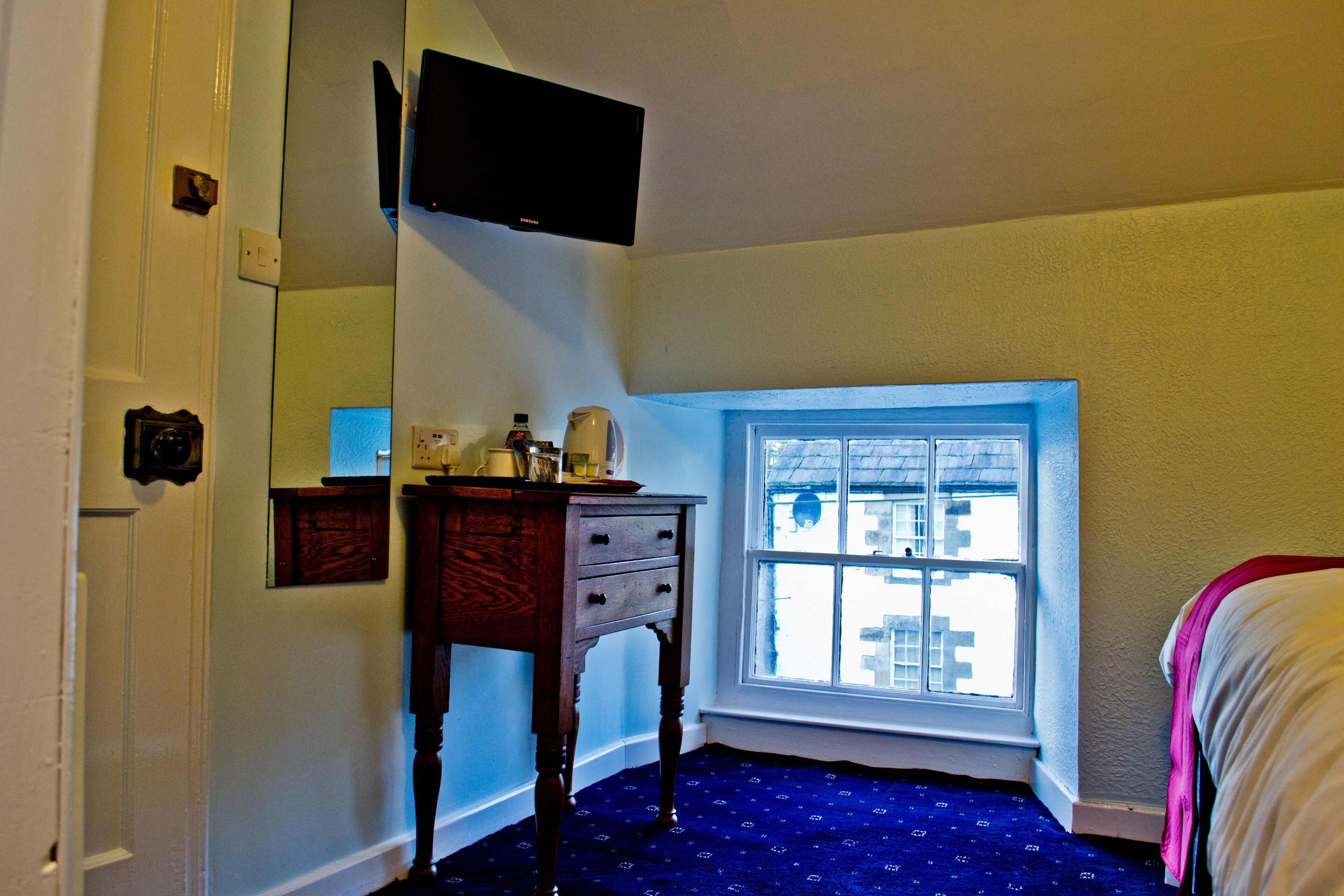 A room with a window, a wooden dresser, a flat-screen TV mounted on the wall, and a bed with a white blanket.