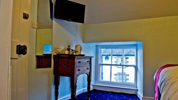 A room with a window, a wooden dresser, a flat-screen TV mounted on the wall, and a bed with a white blanket.