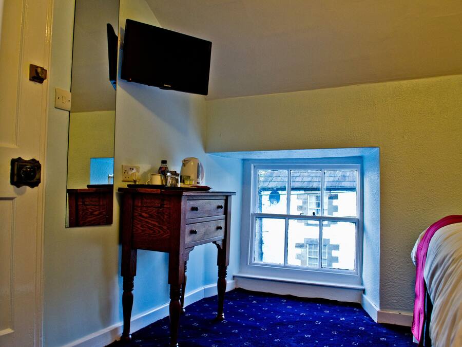 A room with a window, a wooden dresser, a flat-screen TV mounted on the wall, and a bed with a white blanket.