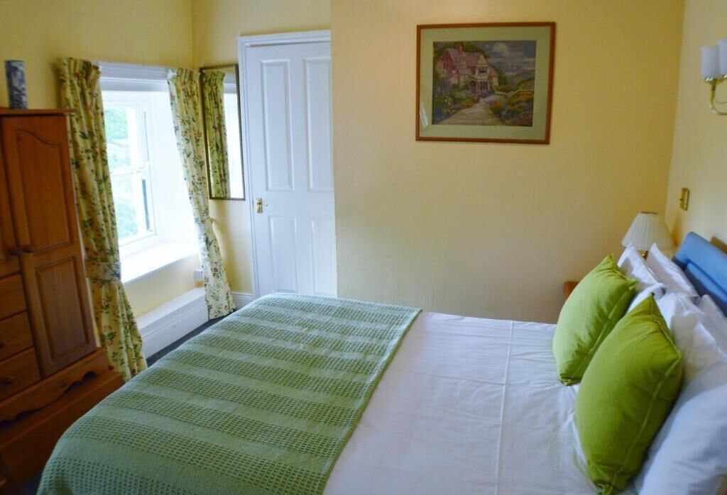 A bedroom with a bed, green pillows, a wooden dresser, a painting on the wall, and a window with curtains.