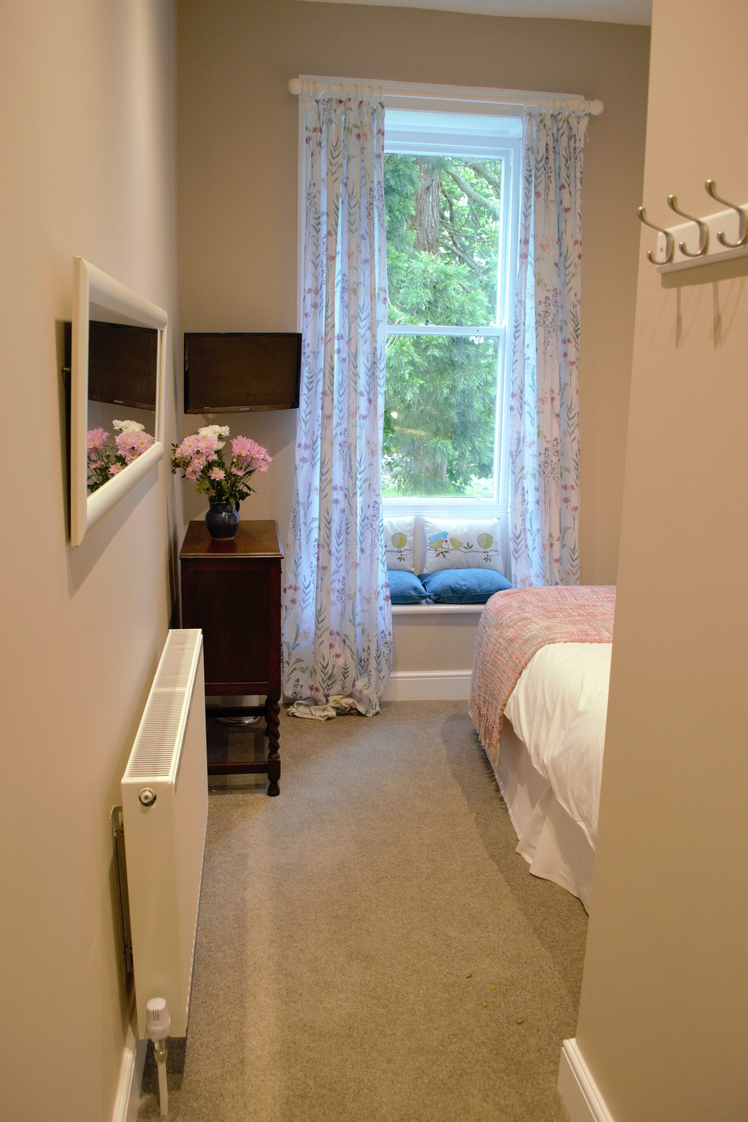 Una habitación estrecha con una cama, una ventana con cortinas florales, un radiador y una pequeña mesa con un florero.