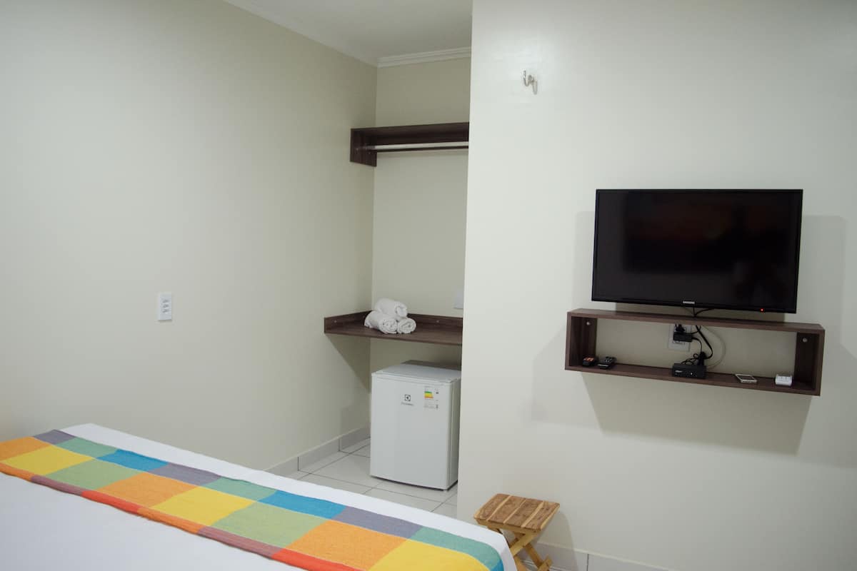 A hotel room with a bed, a small refrigerator, a television mounted on the wall, and a wooden shelf with towels.