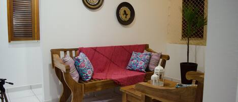 A wooden sofa with colorful cushions, a small wooden table, and a tripod in a room with white walls and a window with blinds.