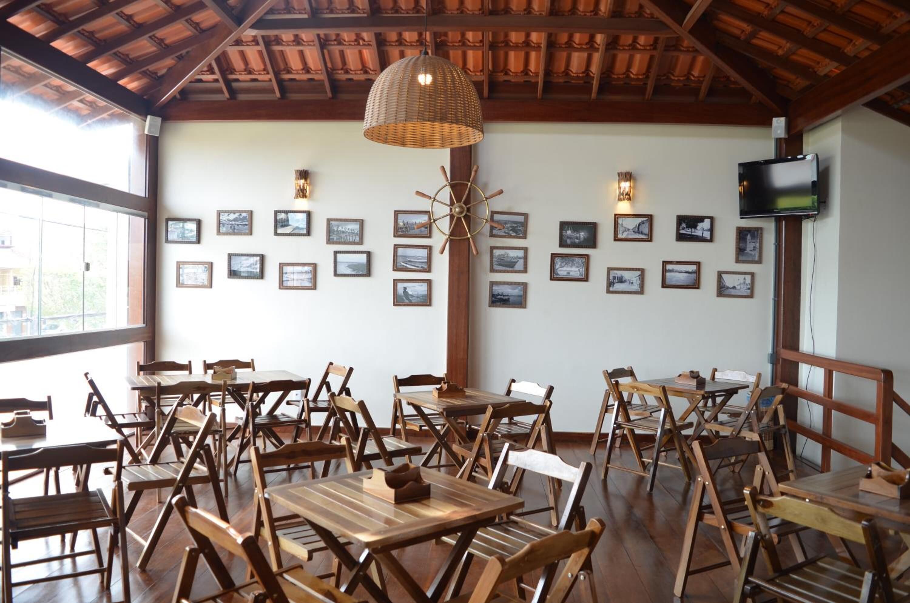 Interior de restaurante com mesas e cadeiras de madeira, uma luminária trançada grande, molduras de quadros penduradas na parede e uma televisão.