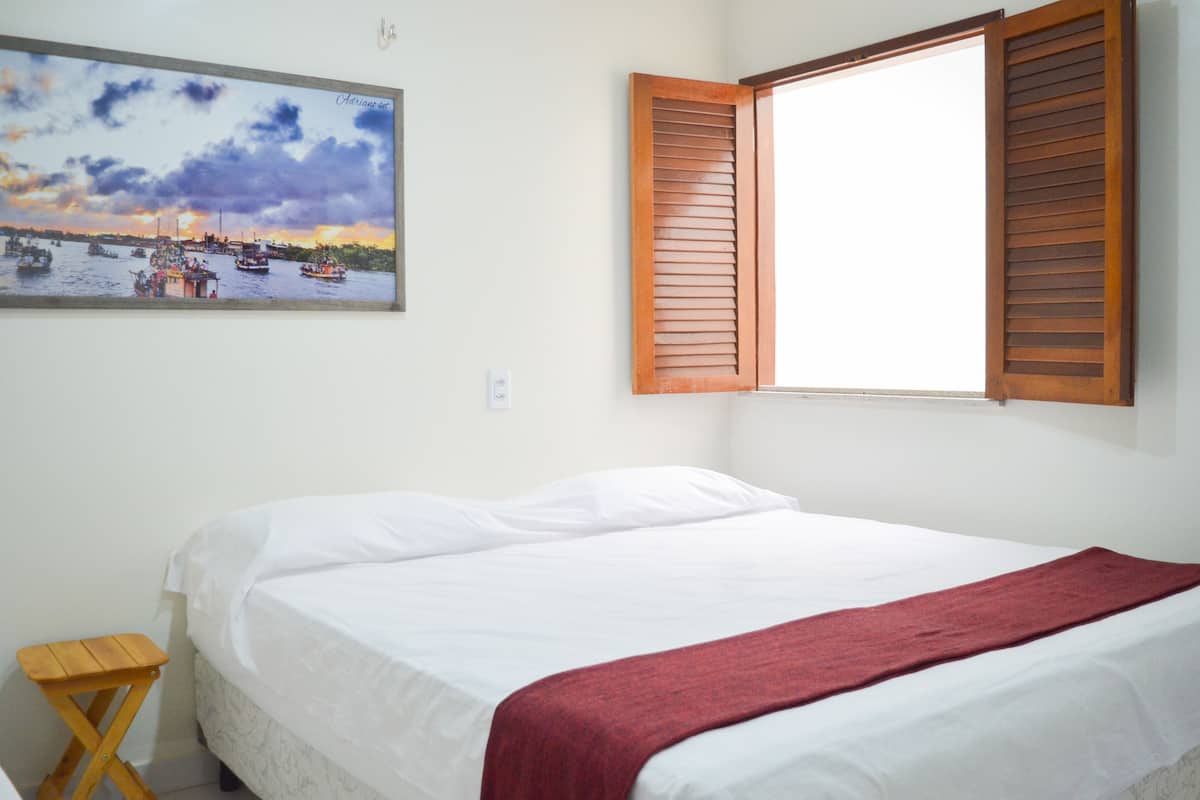 A bedroom with a bed, a window with wooden shutters, a small wooden stool, and a painting of a sunset over a harbor.