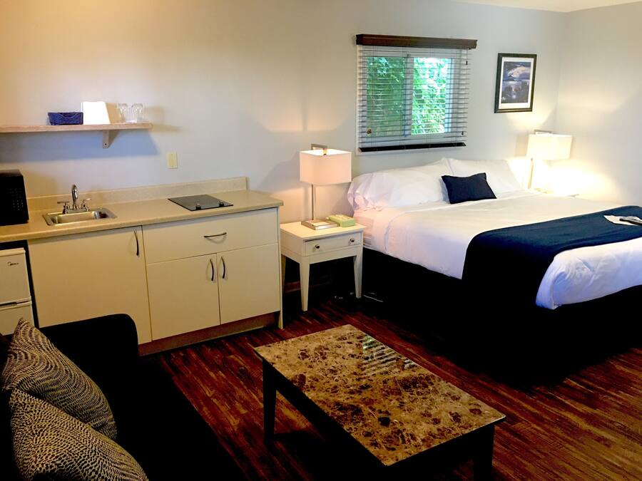 A hotel room with a bed, bedside table, lamp, and a small kitchenette with a sink and cabinets.