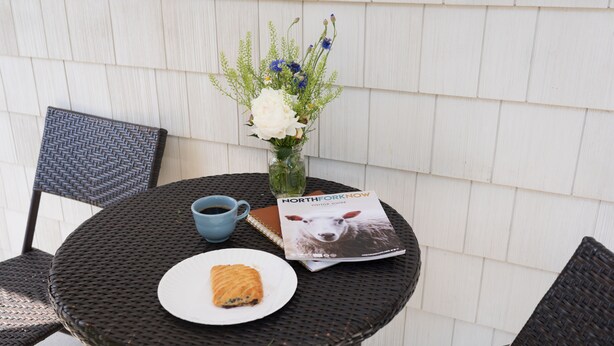 Una mesa redonda con una revista, un plato con un pastel, una taza y un florero con flores.