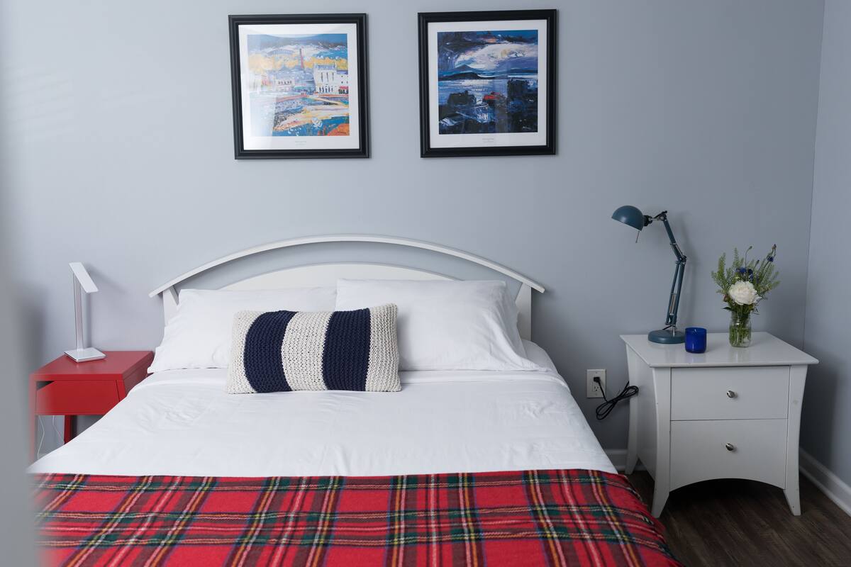 A neatly made bed with a plaid blanket, a white bedside table with a lamp, and two framed artworks on the wall.