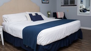 A neatly made bed with a white and navy blue bedspread, a wooden nightstand, a vase with flowers, and a mirror on the wall.