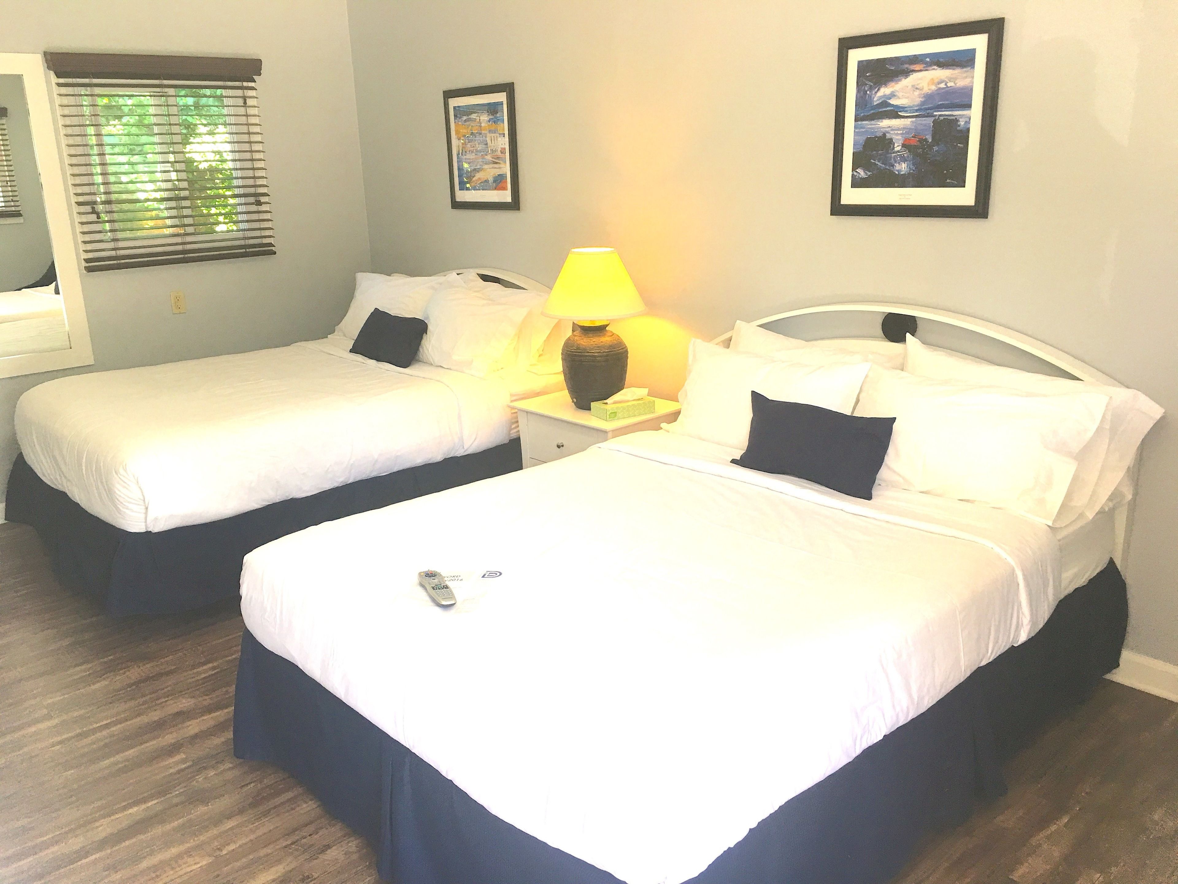 Una camera d'albergo con due letti, un comodino, una lampada e due quadri appesi al muro.