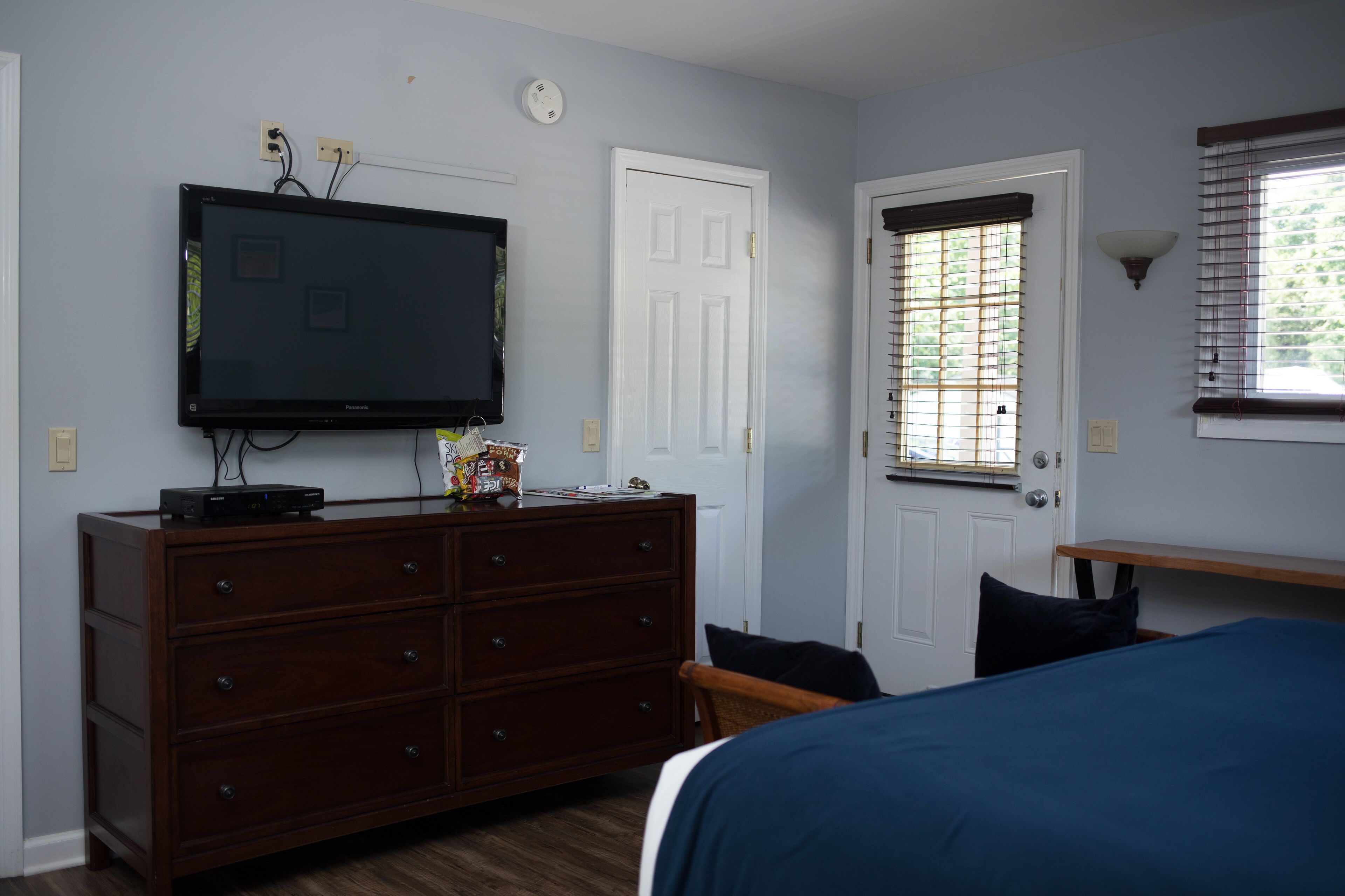 A bedroom with a TV mounted on the wall, a dresser, a door with a window, and a bedside table.