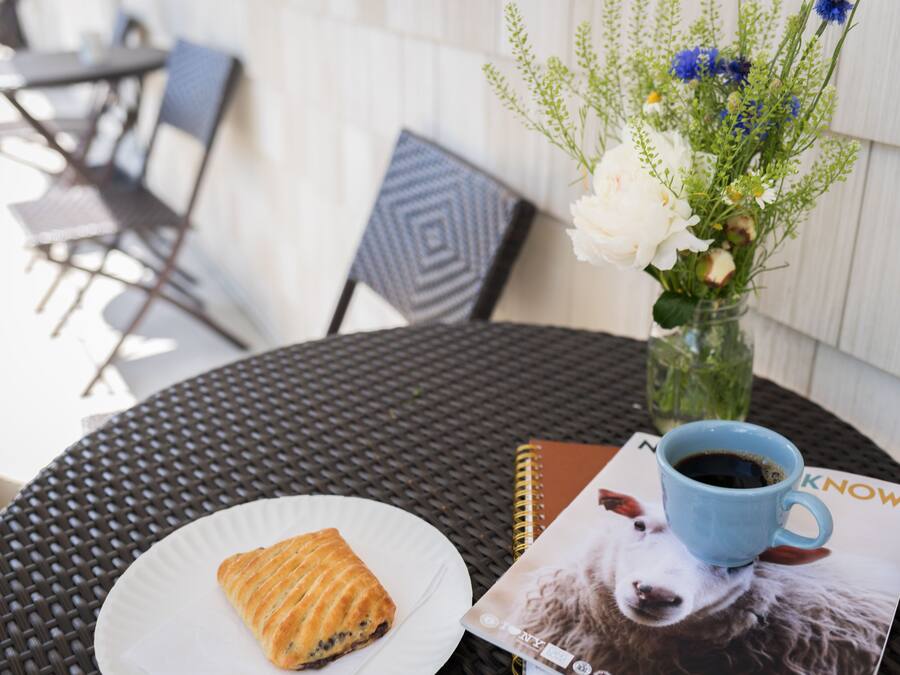 Una mesa redonda con mantel tejido negro, un plato blanco con un pastel, una taza de café azul, un jarrón con flores y una revista con la imagen de un perro.