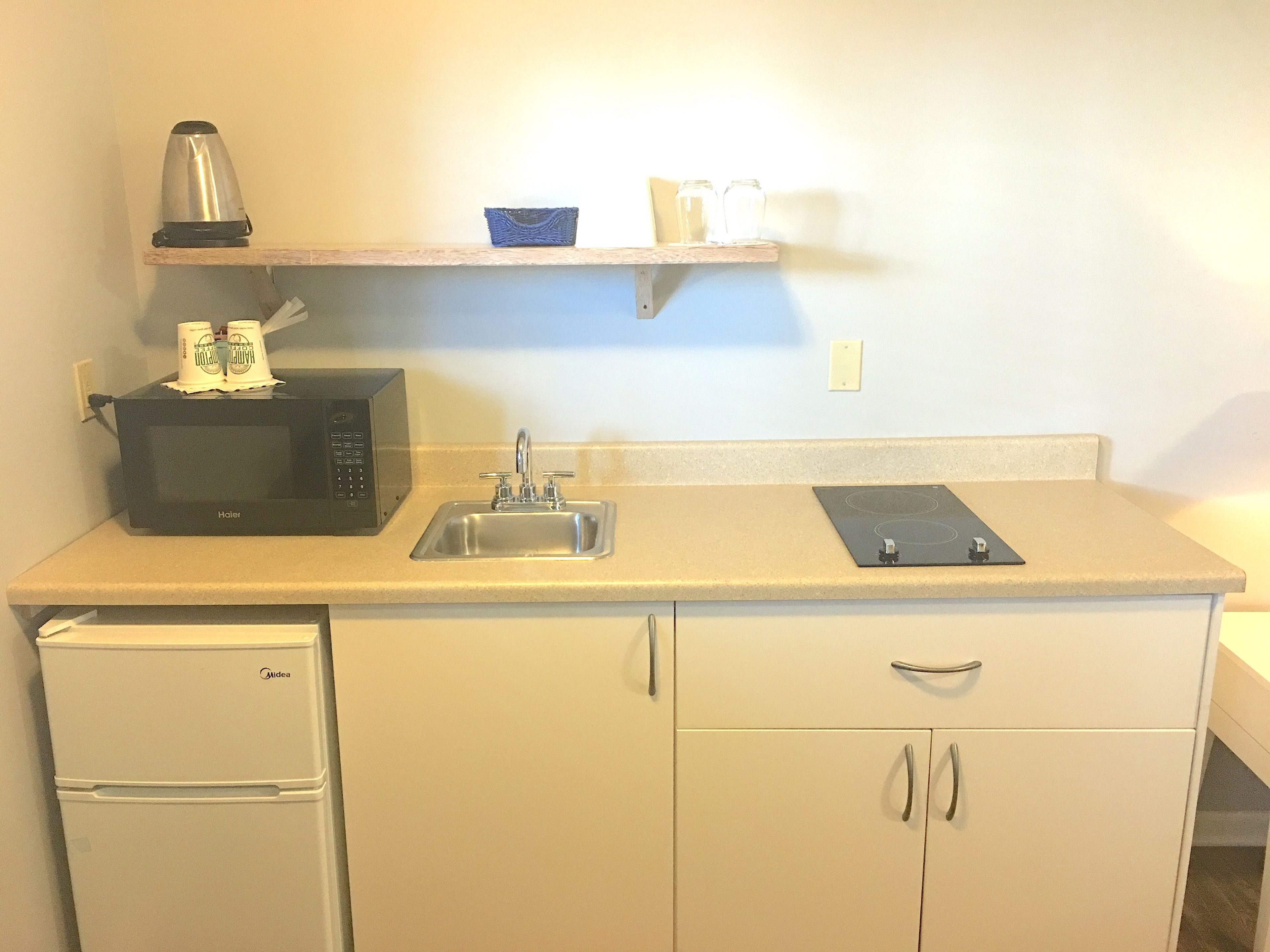 Una kitchenette compacta con microondas, hervidor y un estante pequeño con tazas y un tazón.