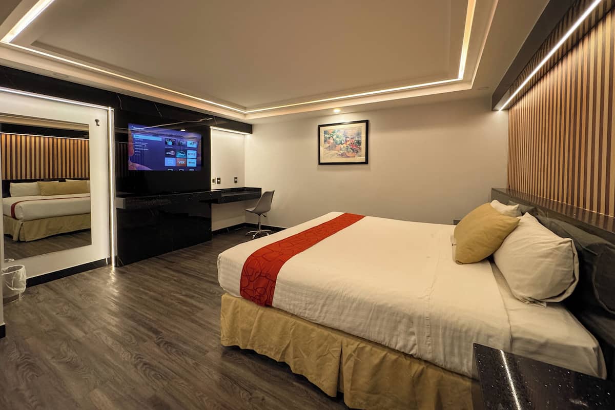 A modern hotel room with a bed, a desk with a computer, a TV, and a mirror.