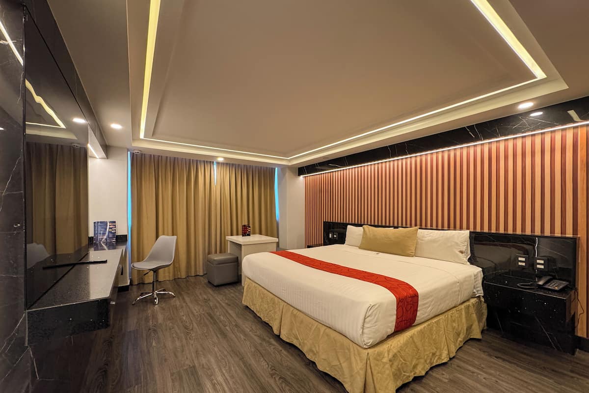 A modern hotel room with a large bed, a desk, a chair, a small table, and a nightstand with a phone.