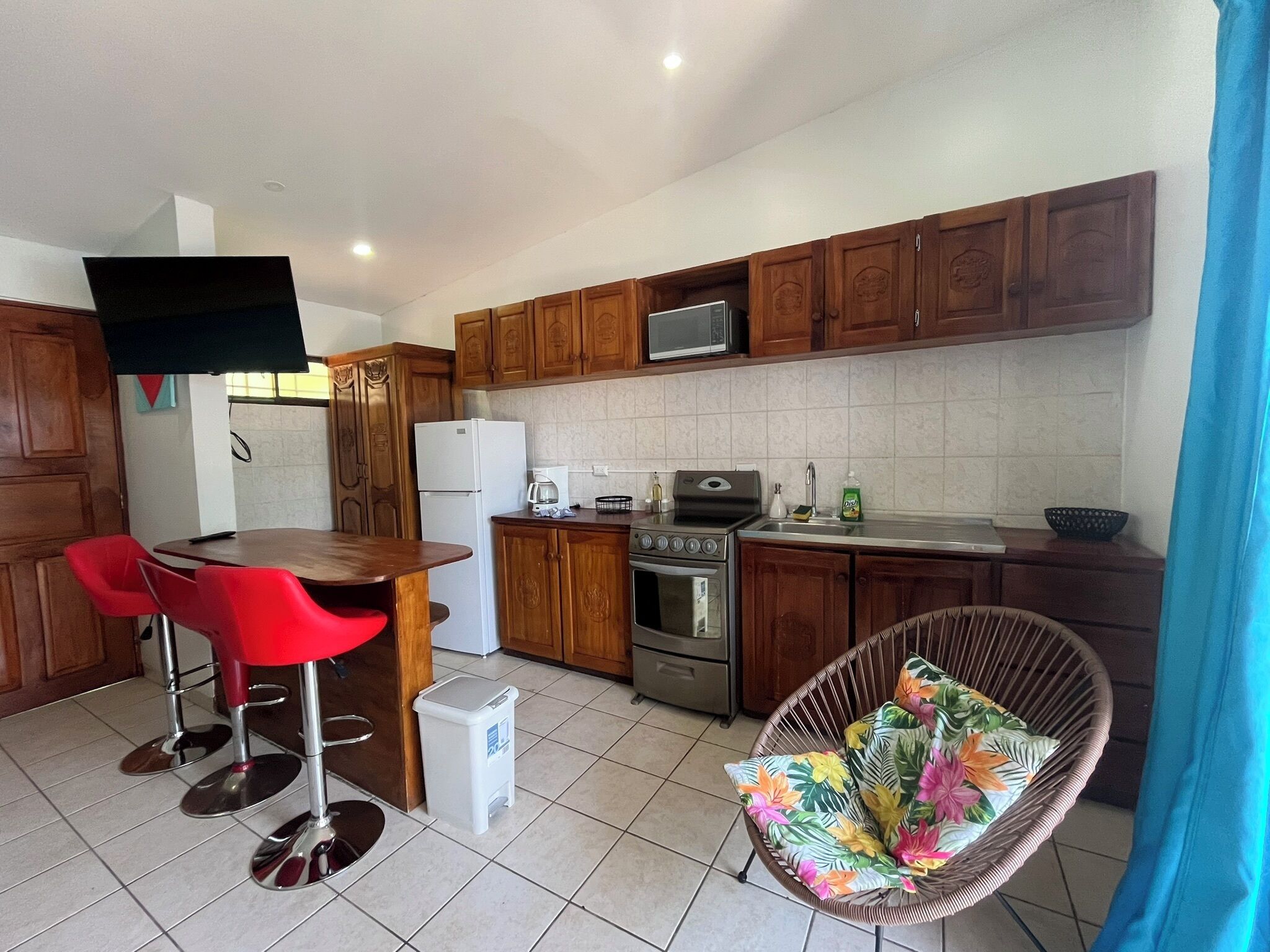 A compact kitchen with wooden cabinets, a microwave, a refrigerator, a stove, and a sink. There are two red bar stools and a wicker chair with a floral cushion.