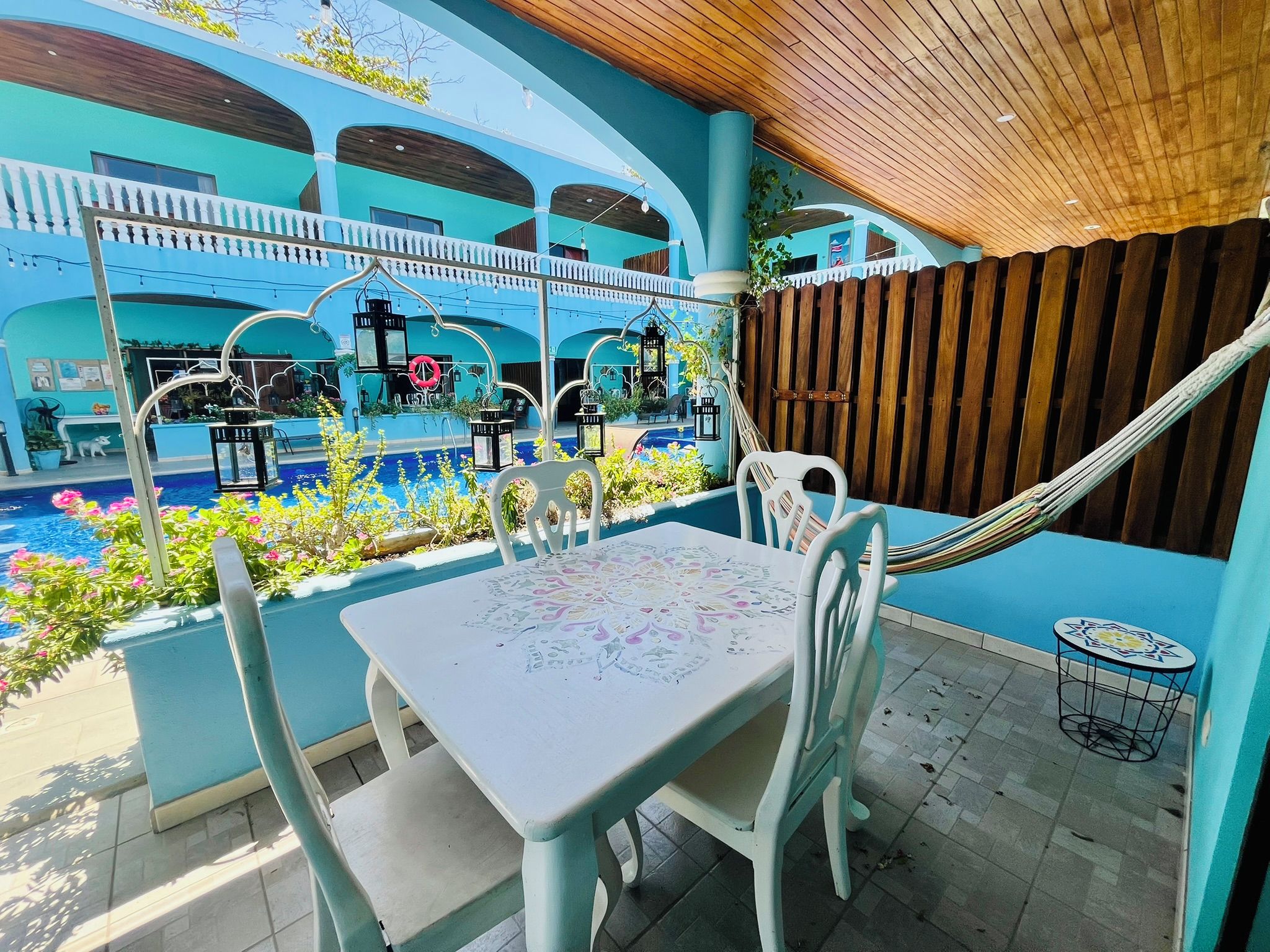 Uma área de estar externa com mesa, cadeiras, uma rede e uma piscina ao fundo.
