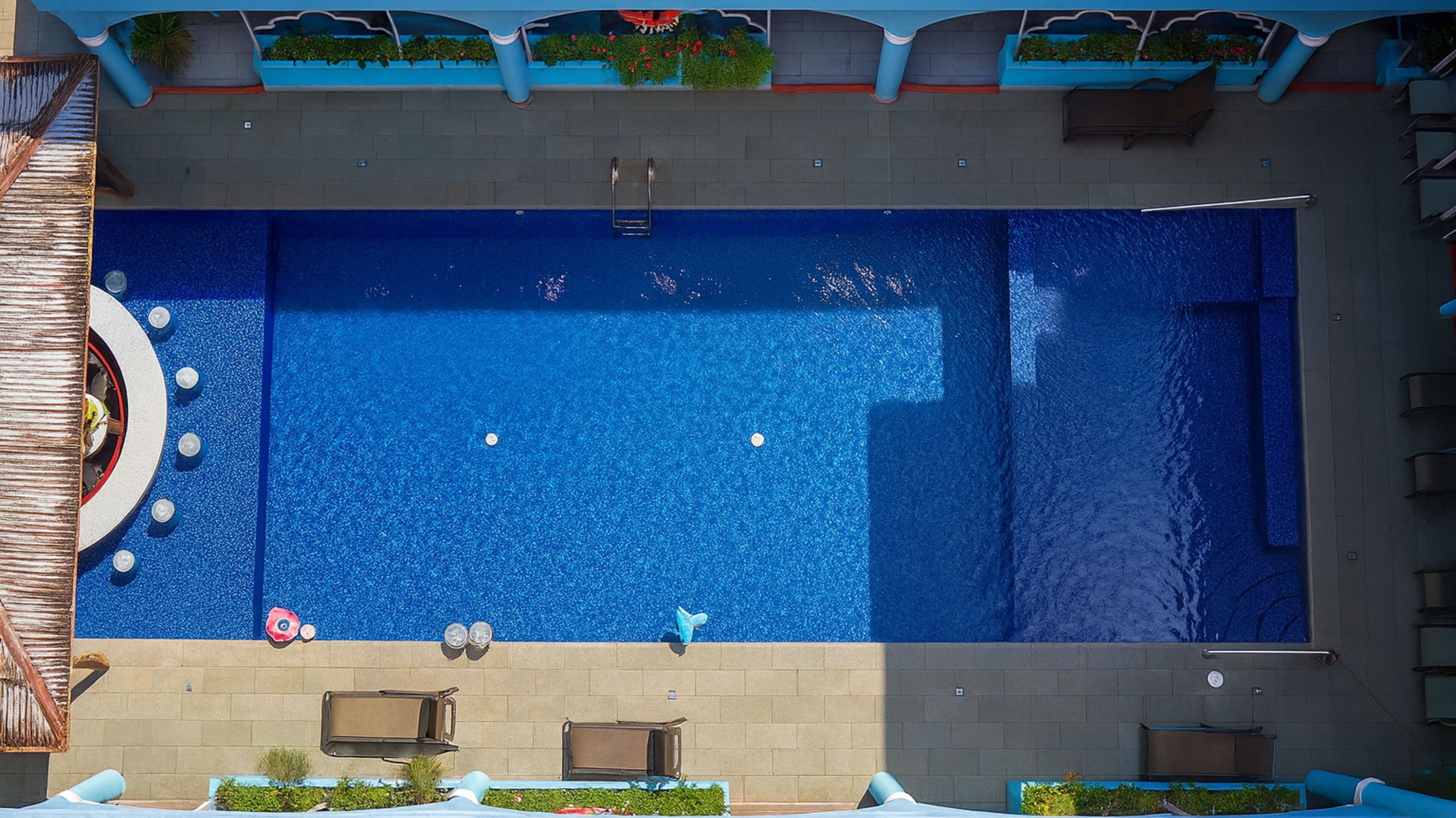 Ein rechteckiger Swimmingpool mit blauer Innenseite, umgeben von einer gefliesten Terrasse und einer niedrigen Mauer.