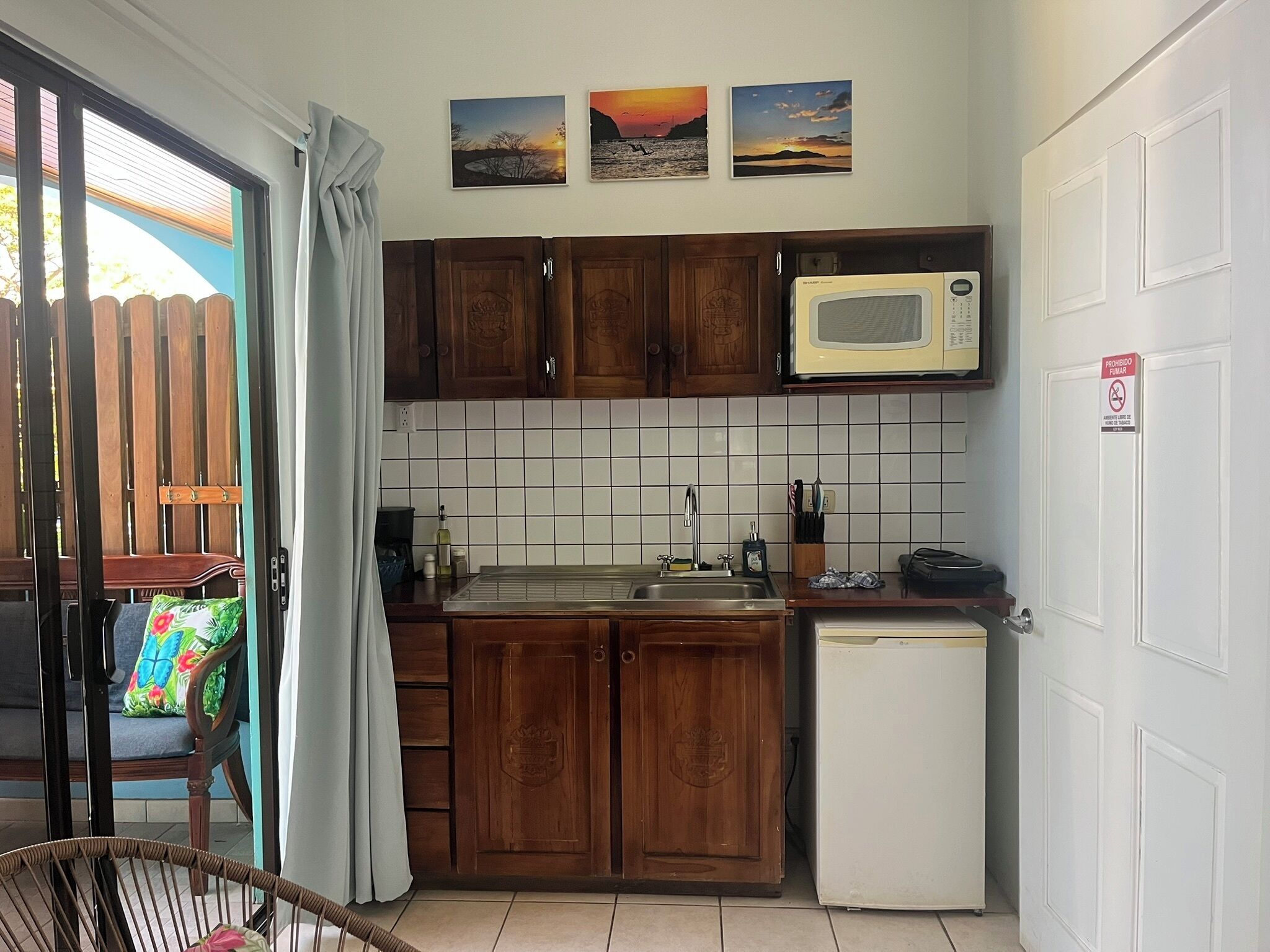 Een compacte keuken met houten kastjes, een magnetron, een spoelbak en een koelkast. Daarboven hangen drie ingekaderde foto’s aan de muur.