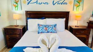 Habitación de hotel con cama, dos mesitas de noche, colcha azul, toallas blancas dispuestas en forma de cisne y almohada colorida.