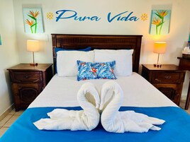 Habitación de hotel con cama, dos mesitas de noche, colcha azul, toallas blancas dispuestas en forma de cisne y almohada colorida.