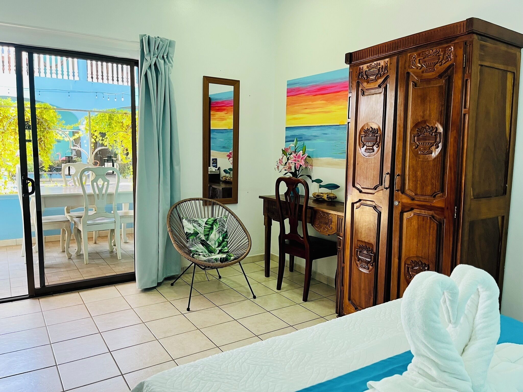 A hotel room with a bed, a desk, a chair, a wardrobe, a sliding glass door, and a painting of a sunset.