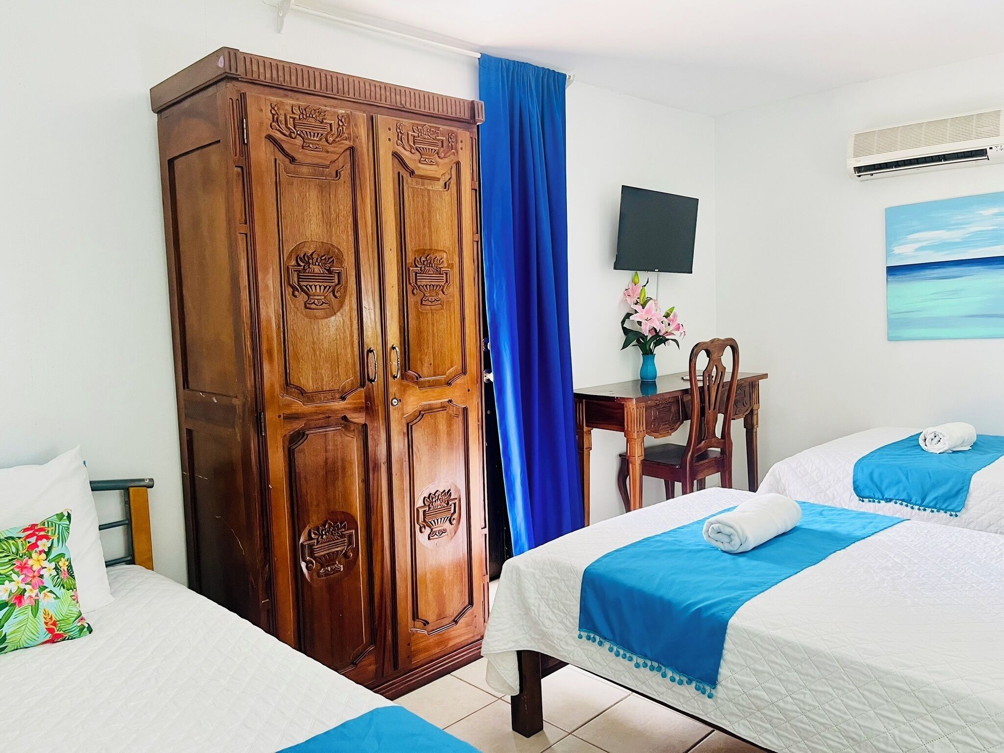 A hotel room with two beds, a wooden wardrobe, a desk with a chair, a TV, and a painting on the wall.