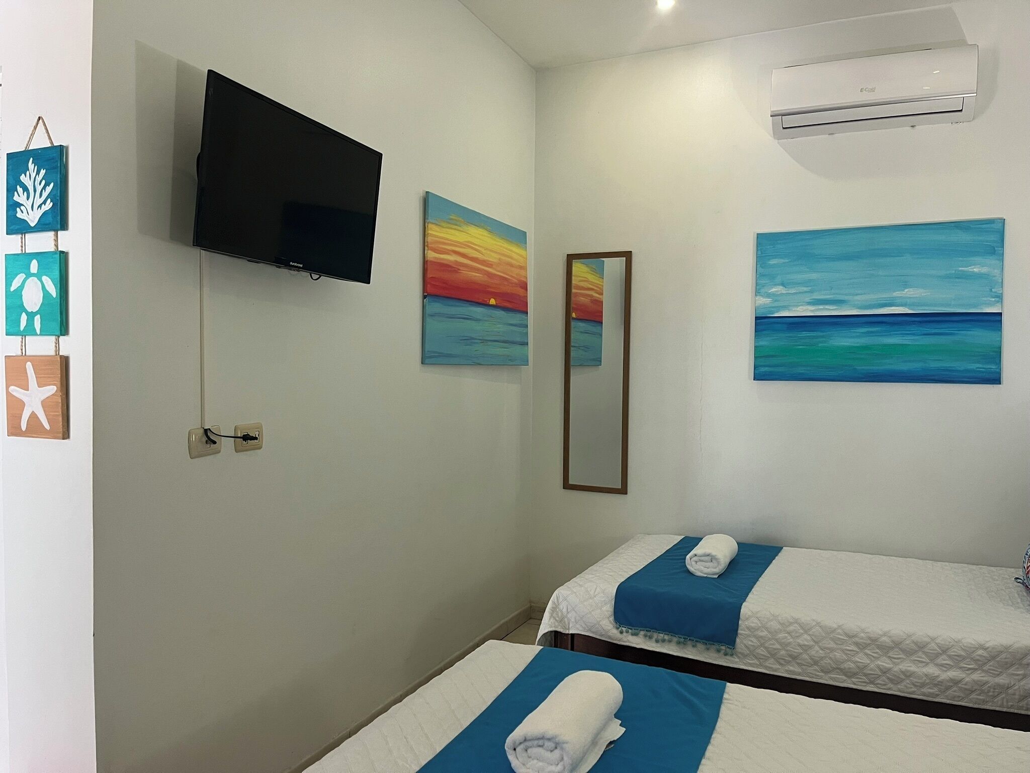 A hotel room with two beds, a flat-screen TV mounted on the wall, and two paintings depicting seascapes.