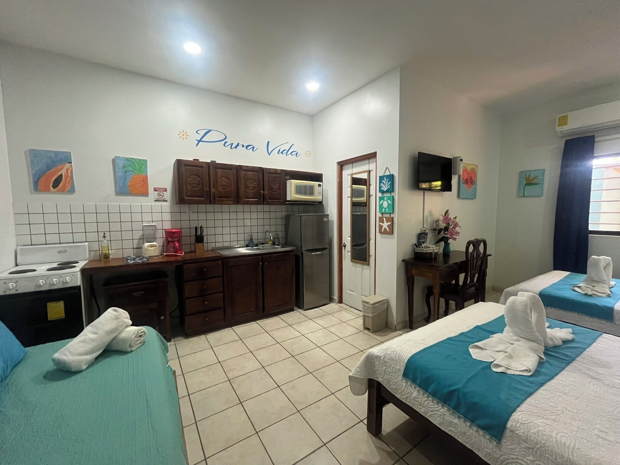 A compact hotel room with a kitchenette, a bed with a blue bedspread, and a small sitting area with a table and chair.