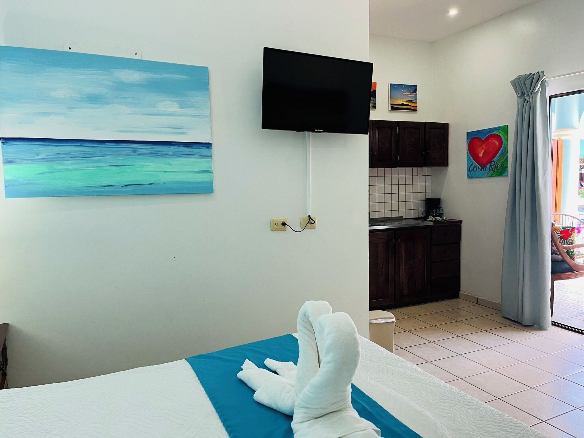 A hotel room with a bed, a painting of a beach, a television, a kitchen area, and a window with curtains.