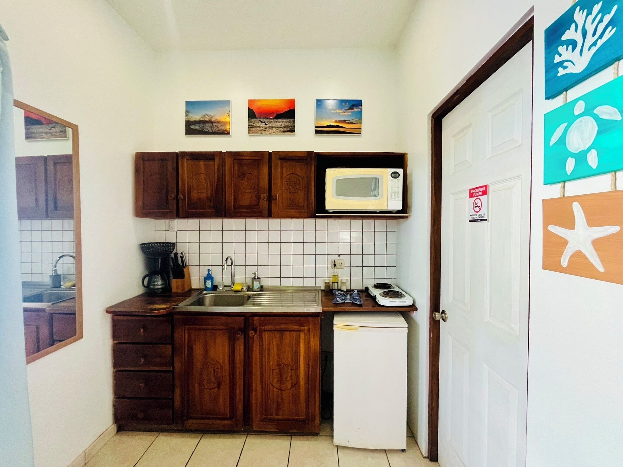A compact kitchen with wooden cabinets, a microwave, a sink, and a small refrigerator. There are three framed pictures on the wall above the cabinets.