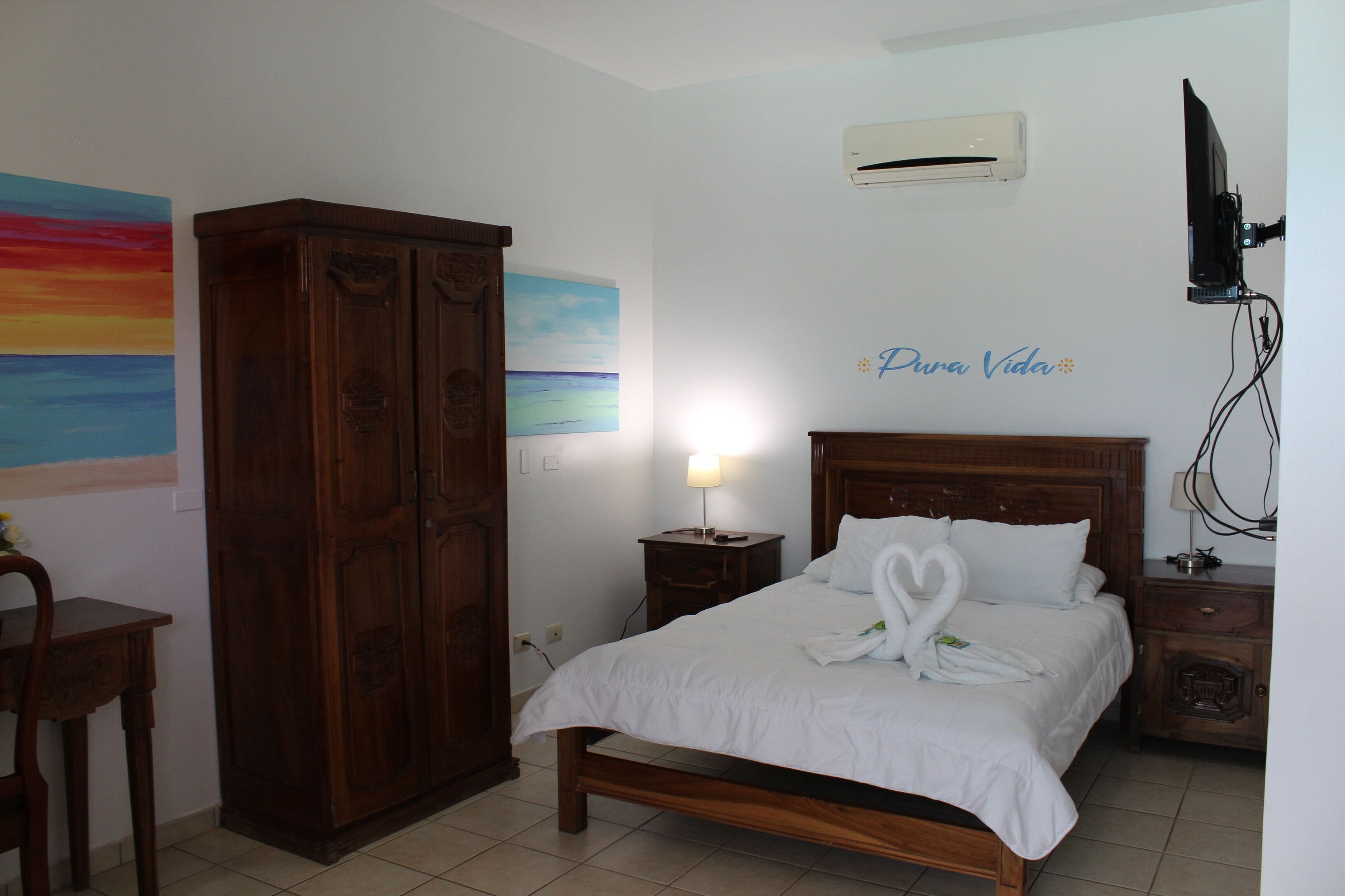 A bedroom with a wooden bed, a nightstand with a lamp, a wardrobe, a painting on the wall, and an air conditioning unit.