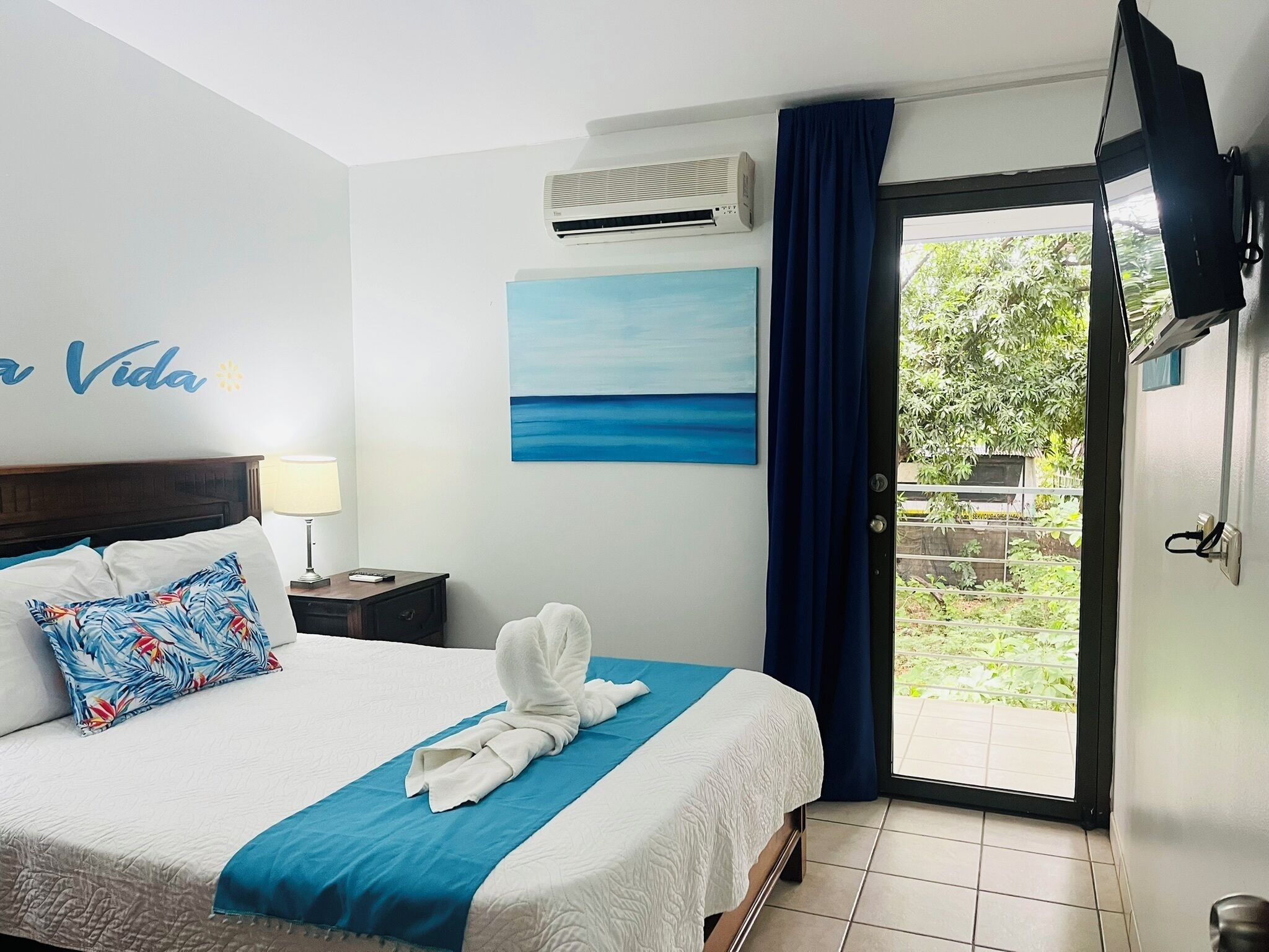 Habitación de hotel con cama, un cuadro, equipo de aire acondicionado, mesita de noche, lámpara y vistas a través de un ventanal.
