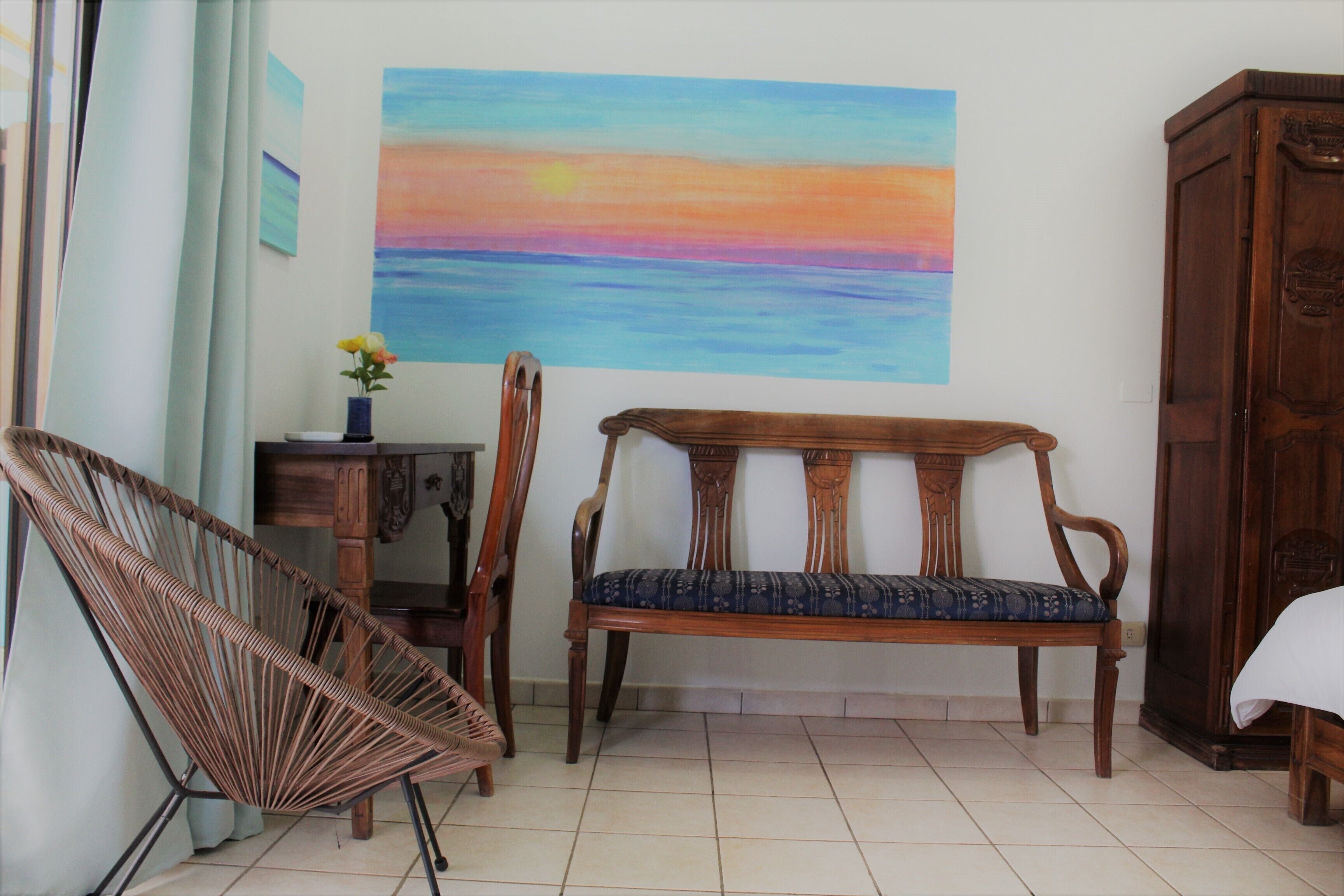 Una habitación con un banco de madera, una silla de mimbre, una mesita con un jarrón de flores y un cuadro grande de un atardecer en la pared.