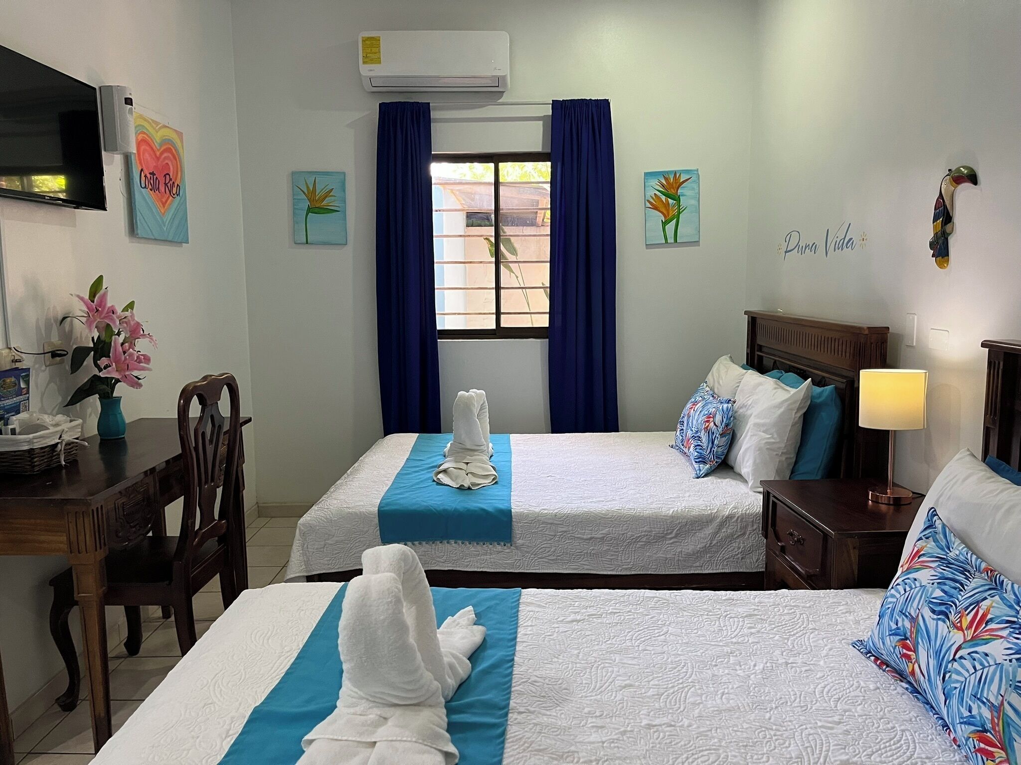 Habitación de hotel con dos camas, un escritorio, una silla, aire acondicionado y obras de arte en las paredes.