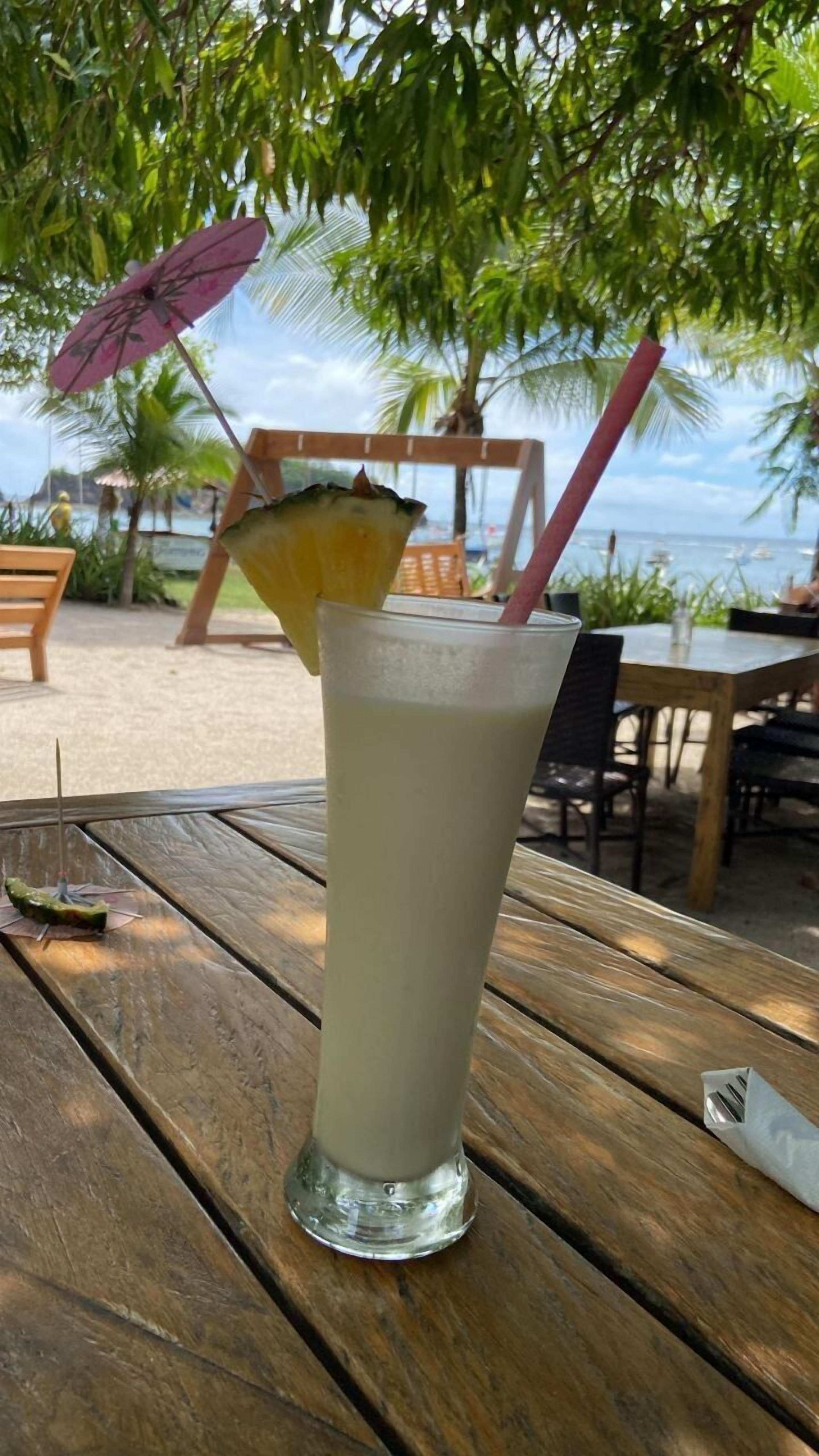 A tall glass with a straw and a pineapple slice on a wooden table at a beachside location.