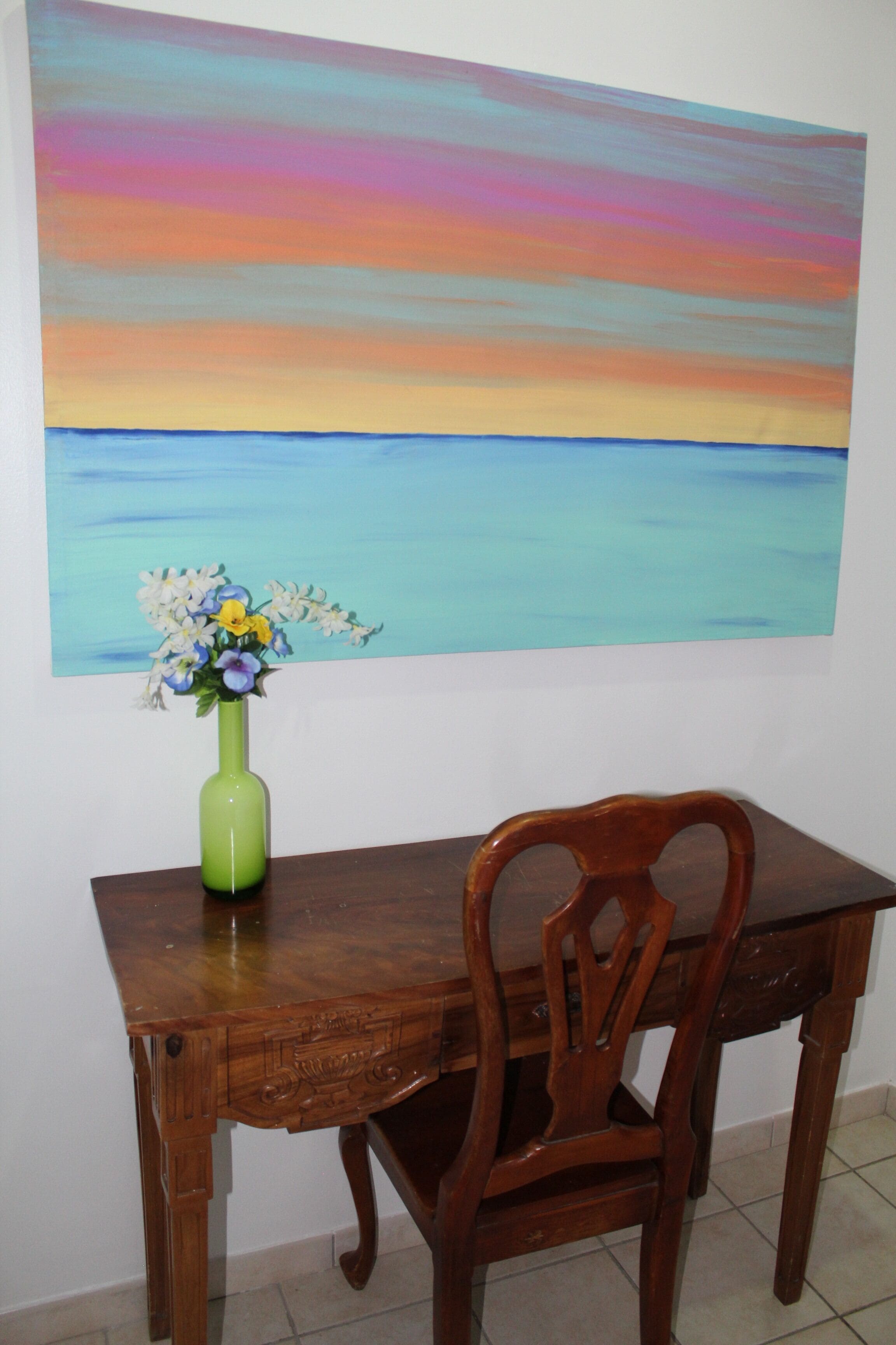 Una mesa de comedor de madera con un jarrón verde lleno de flores, una silla de madera y un cuadro grande de un atardecer sobre el agua en la pared.