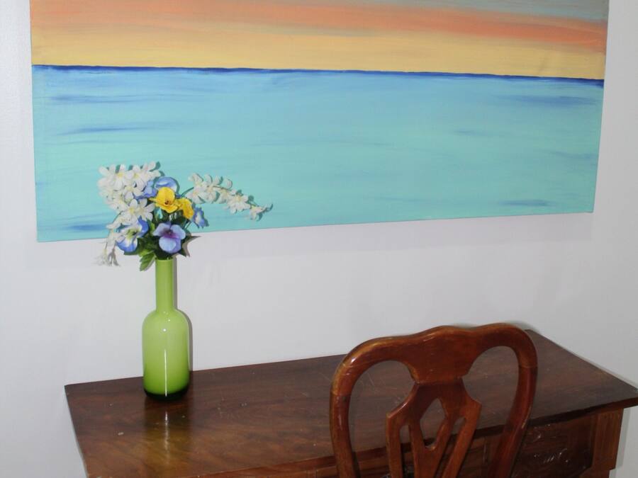 Una mesa de comedor de madera con un jarrón verde lleno de flores, una silla de madera y un cuadro grande de un atardecer sobre el agua en la pared.
