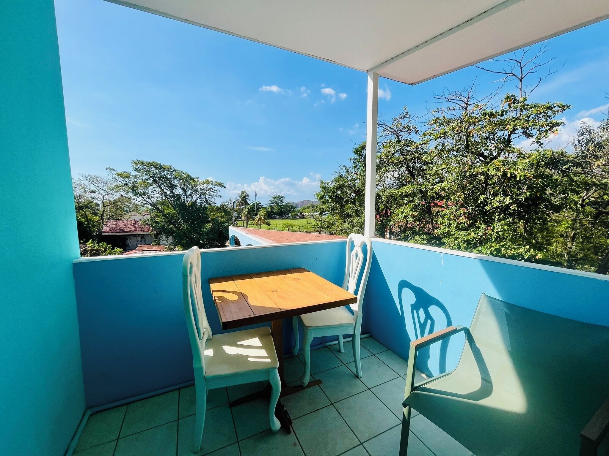 Ein Balkon mit Tisch und Stühlen, Blick auf Grünflächen und Gebäude, und ein klarer Himmel.