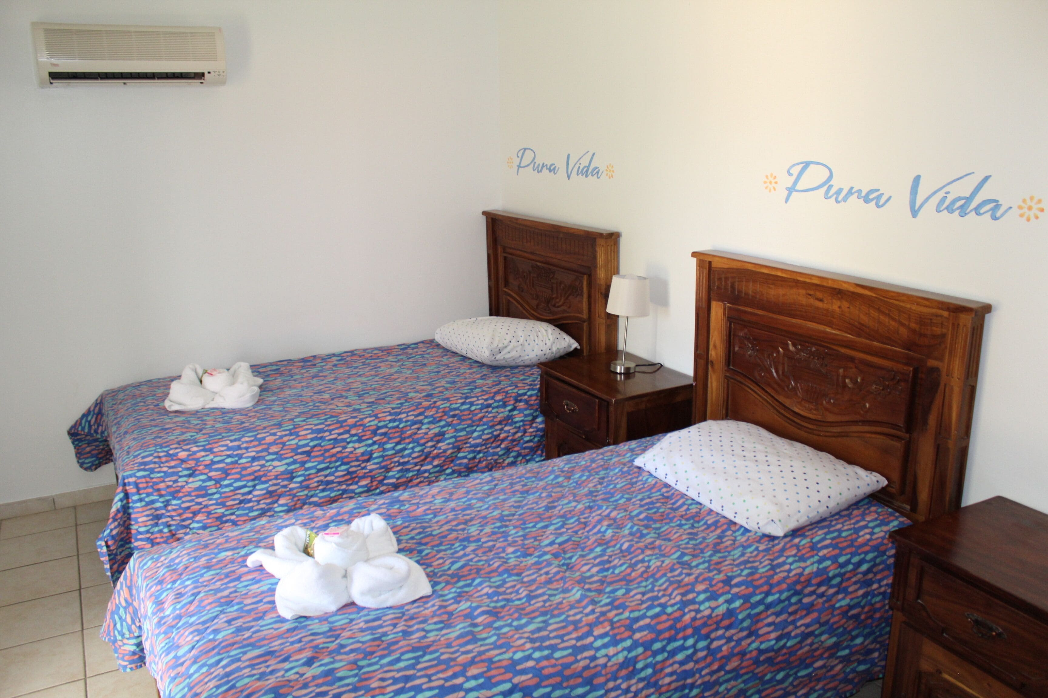 A hotel room with two beds, each with a patterned bedspread and white towels. There is a wooden headboard on each bed, a bedside table with a lamp, and a wall-mounted air conditioner. The room has a tiled floor and a wall with the text "Pura Vida".