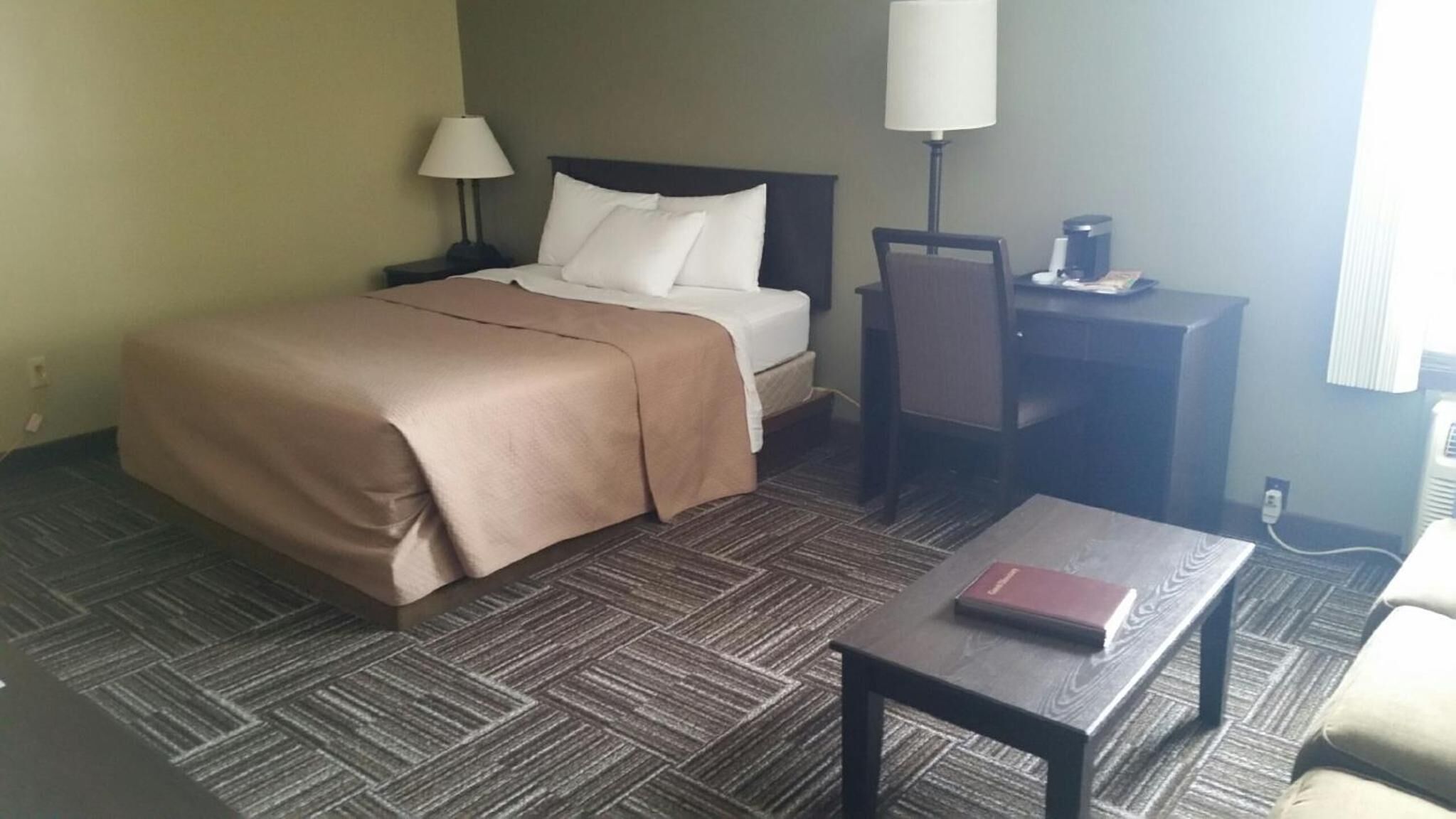 A hotel room with a bed, a desk, a chair, a coffee maker, and a lamp.